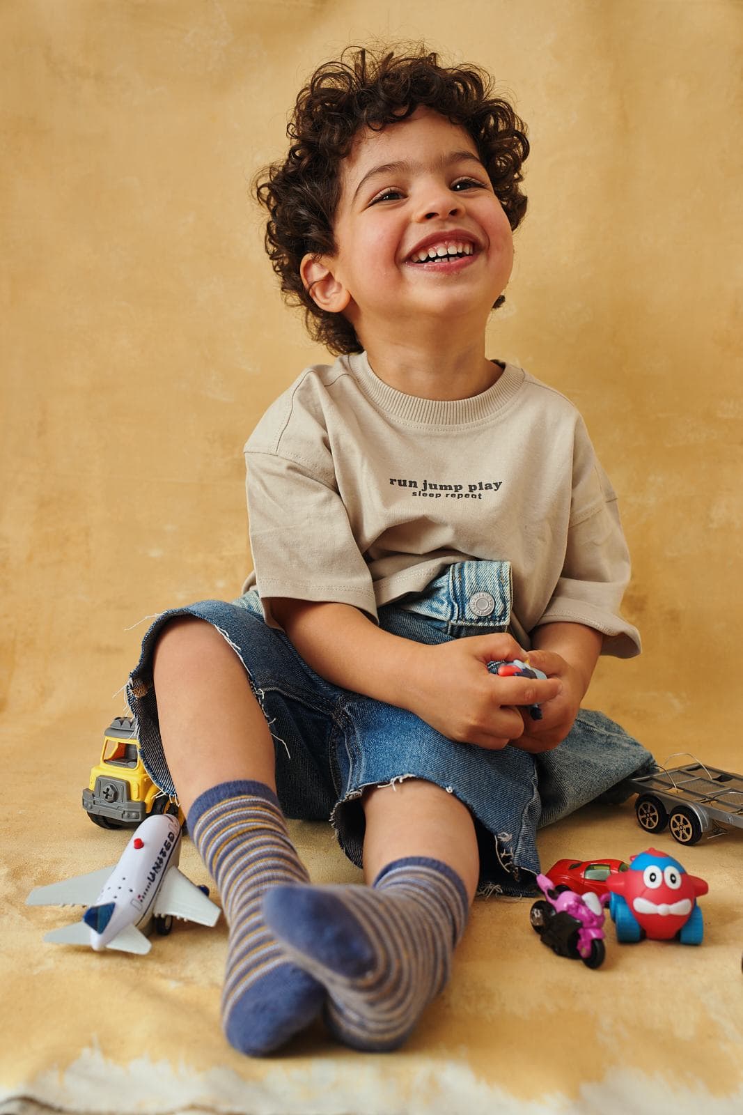 Play-based studio portrait of a toddler laughing among toy cars on a warm ochre backdrop in Vancouver by Una Zhu