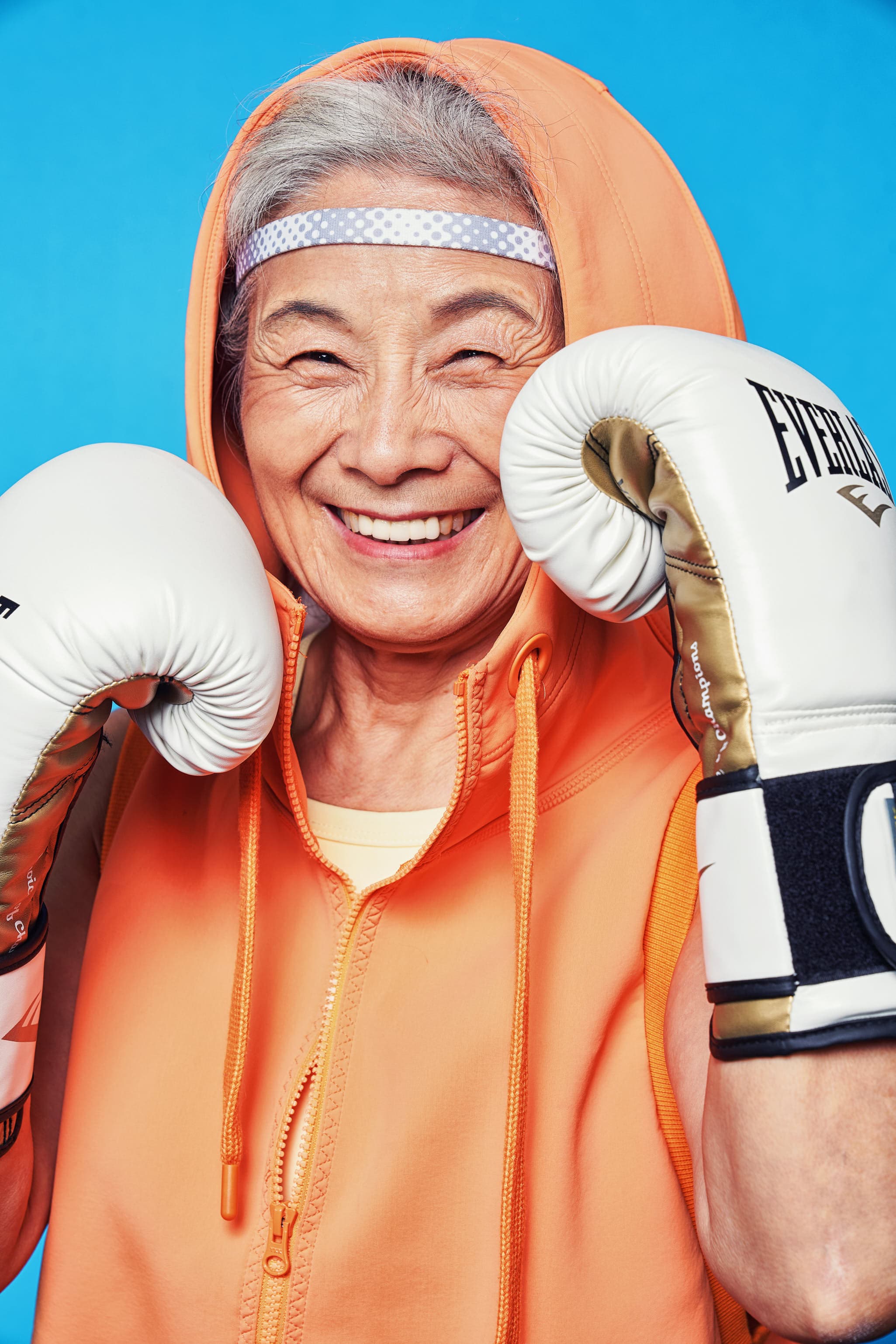 Lifestyle fitness editorial close-up portrait of elderly woman in boxing gloves and orange hoodie by Una Zhu
