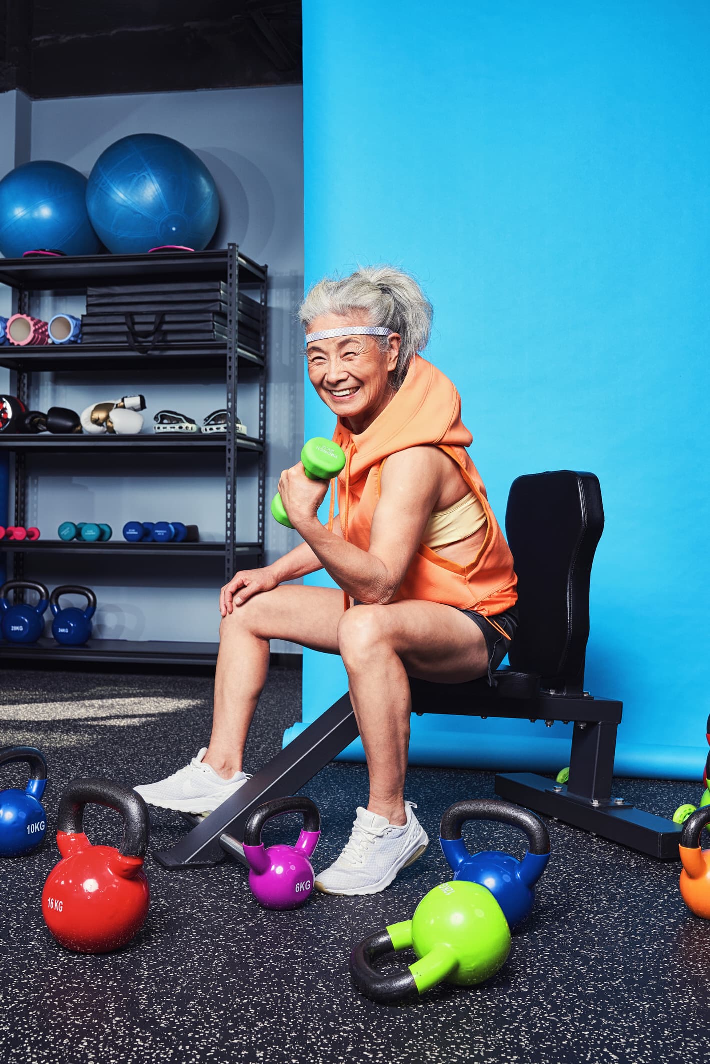 Fitness lifestyle editorial with elderly woman lifting dumbbell at gym with colorful kettlebells by Una Zhu
