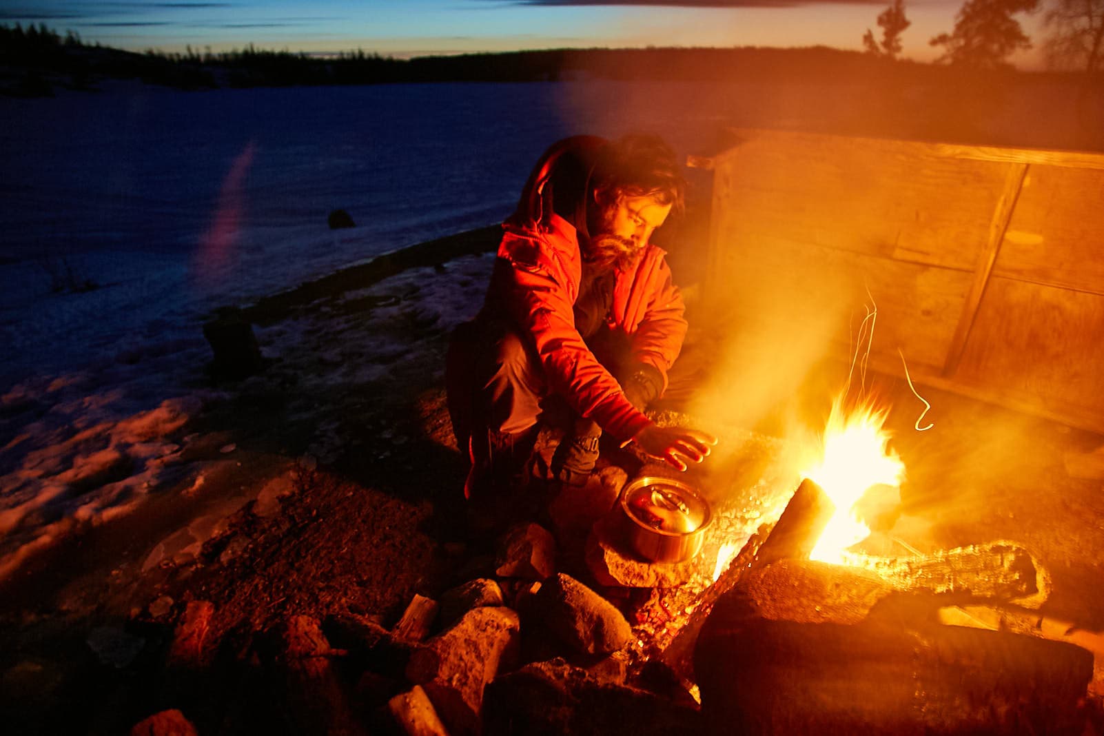 Documentary outdoor lifestyle photography with man cooking by campfire in snowy wilderness at twilight by Una Zhu