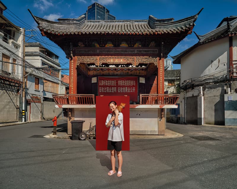 Documentary street portrait of woman holding dog against red backdrop under traditional Shanghai gate by Una Zhu
