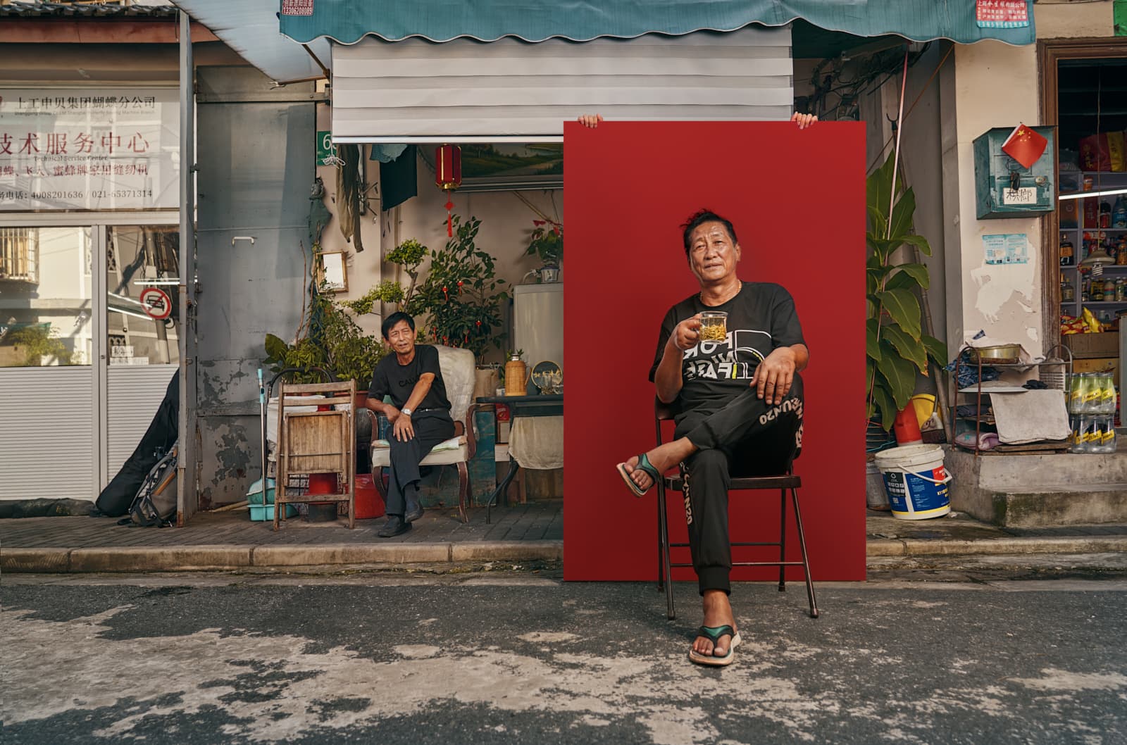 Editorial street portrait of man seated with tea against red backdrop outside Shanghai shopfront by Una Zhu