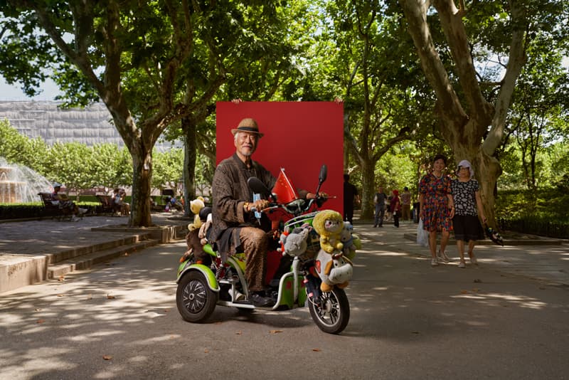 Street portrait of elderly man on decorated tricycle against red backdrop in Shanghai park by Una Zhu