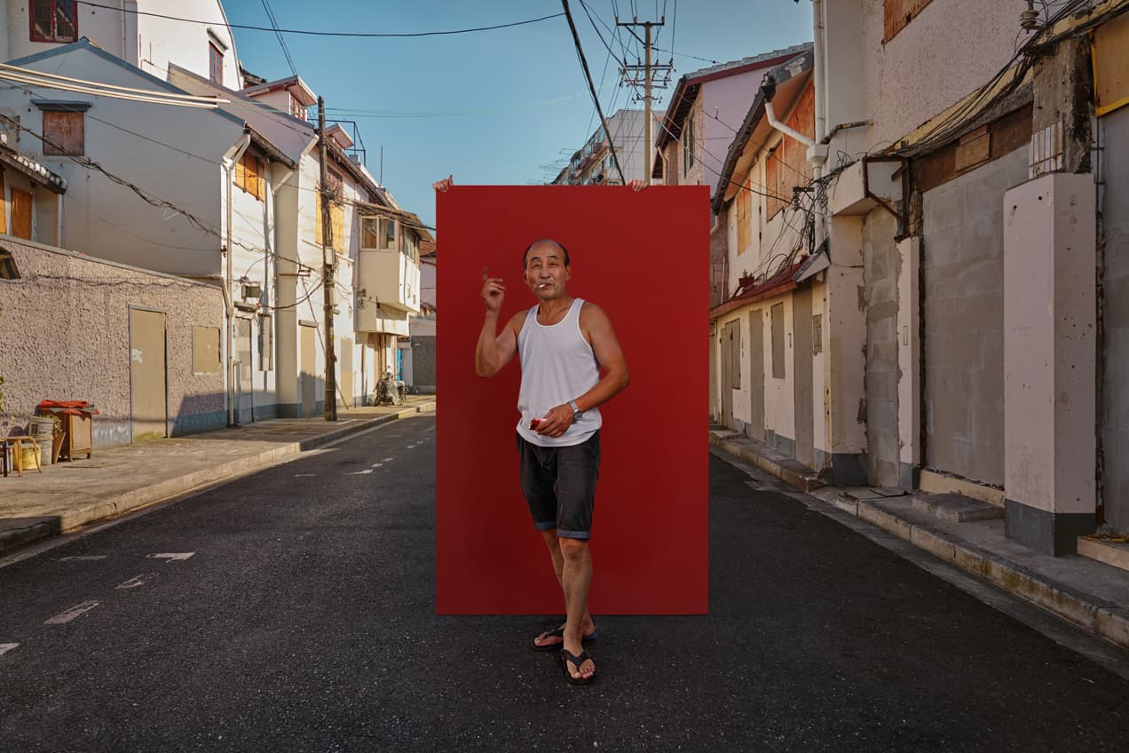 Editorial street portrait of man in white tank top smoking against red backdrop in Shanghai lane by Una Zhu