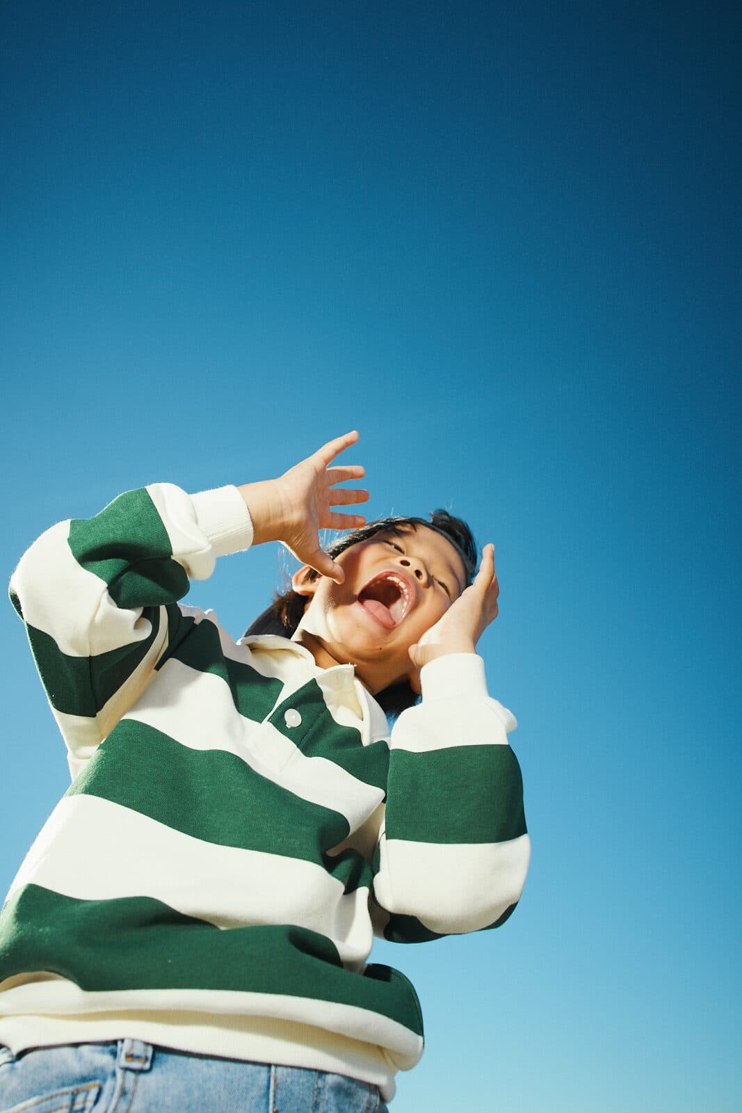 Unposed photo of a child shouting joyfully against a clear blue sky at a Vancouver beach by Una Zhu