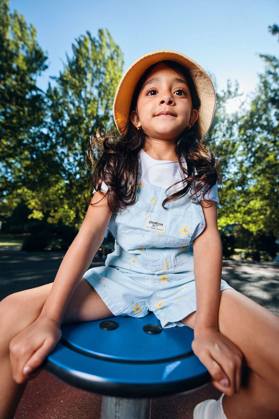 Unposed natural light photo of a girl in an orange sun hat on a playground spinner in Vancouver by Una Zhu
