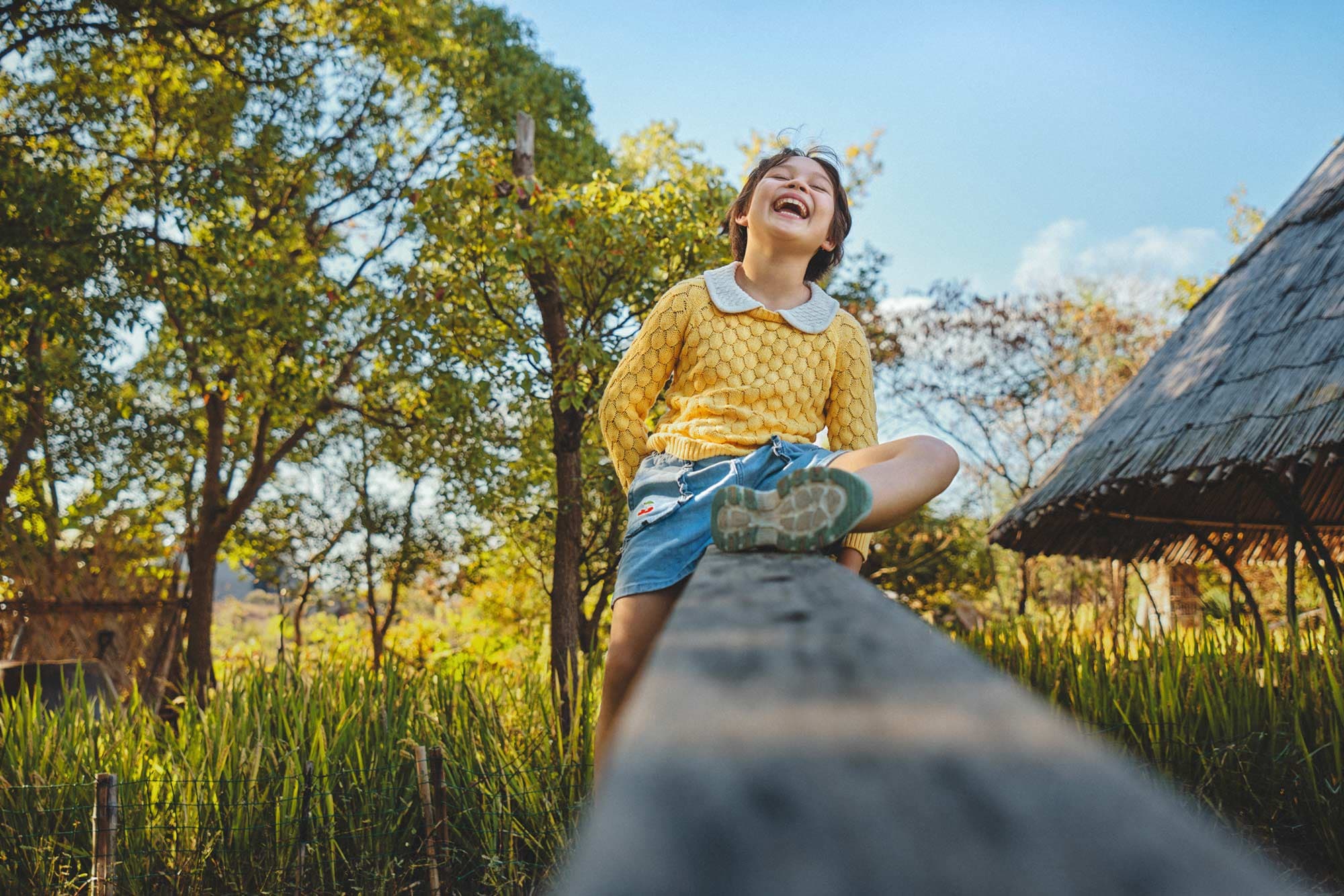 Natural light kids lifestyle photography with child laughing on wooden beam outdoors by Una Zhu