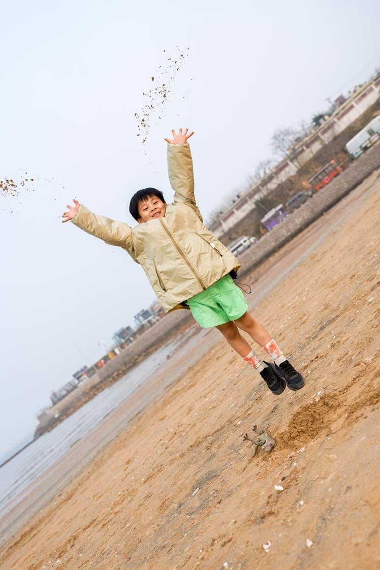 Commercial kids apparel shoot with child jumping on beach in outerwear by Una Zhu