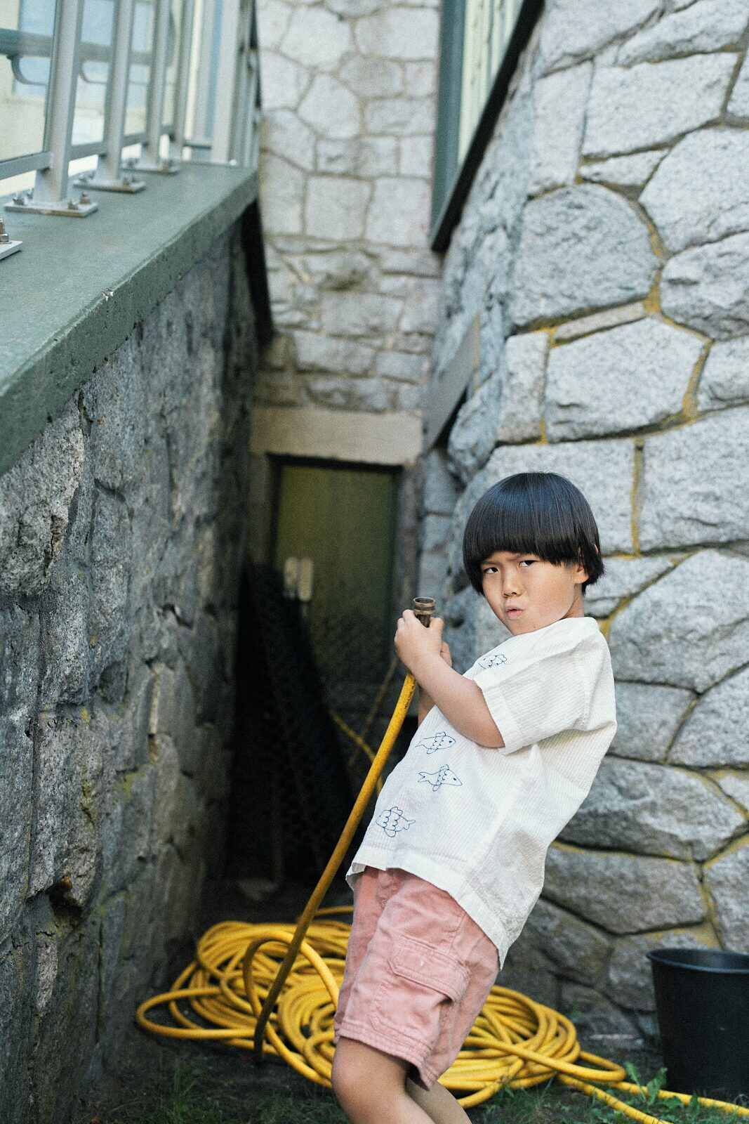 Boy playing with a coiled yellow garden hose beside a stone wall during a candid play session in Vancouver by Una Zhu